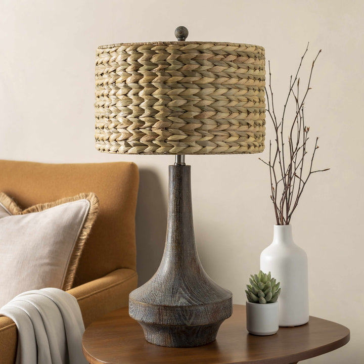 Table lamp with woven shade on a wooden table next to a vase and plant, with a couch in the background.

Kabankalan Seagrass Resin Table Lamp